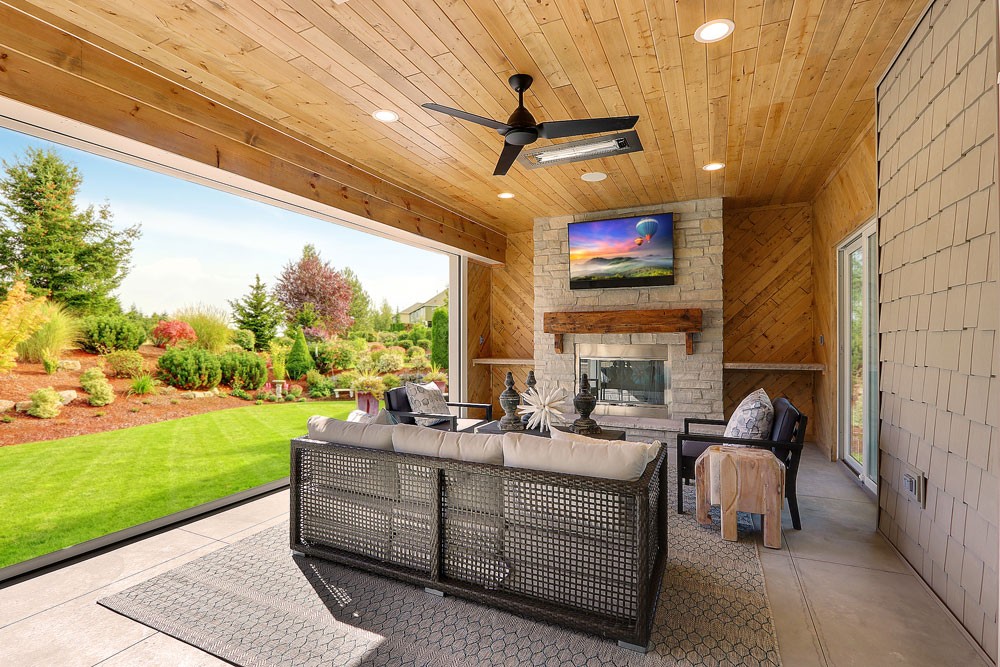 Covered outdoor kitchen and bar area with ceiling fans, chandeliers, built-in grill, and mounted TVs overlooking a green wooded backyard.