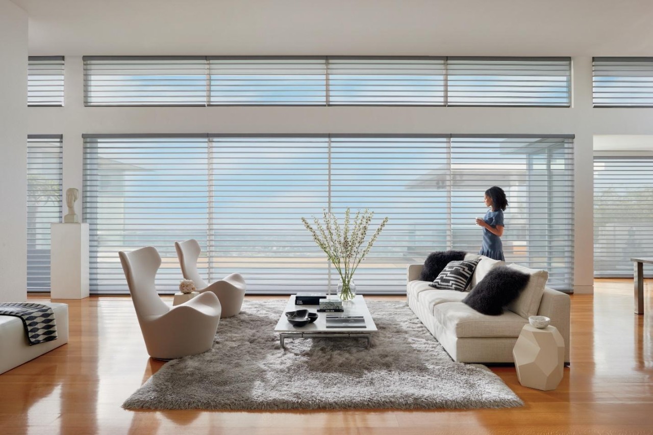 A woman standing in front of large picturesque windows with sheer blinds lowered.