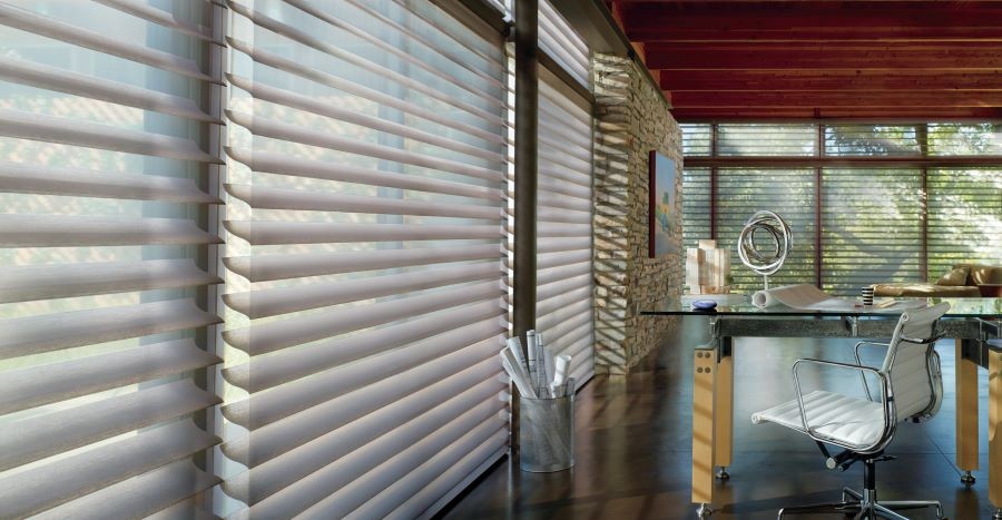Hunter Douglas motorized window blinds lowered over floor-to-ceiling windows in a home office.