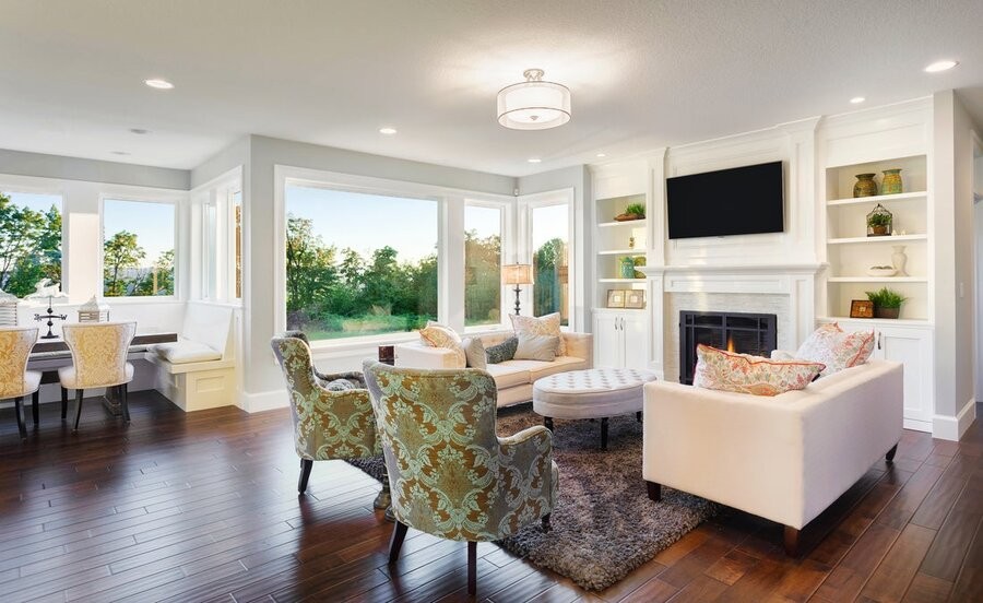 An open living room space featuring seating, a mounted TV above a fireplace, and windows in the background.