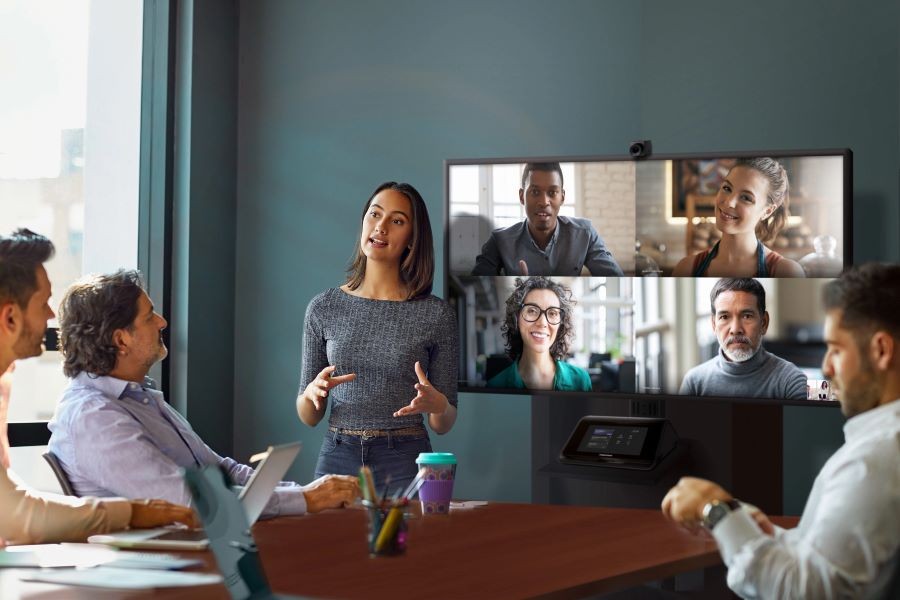 A conference room with a display with four virtual participants, a speaker, and people sitting at a table.